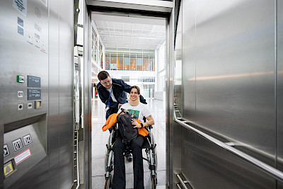Ein Service Agent schiebt eine junge Frau im Rollstuhl in einen Fahrstuhl.