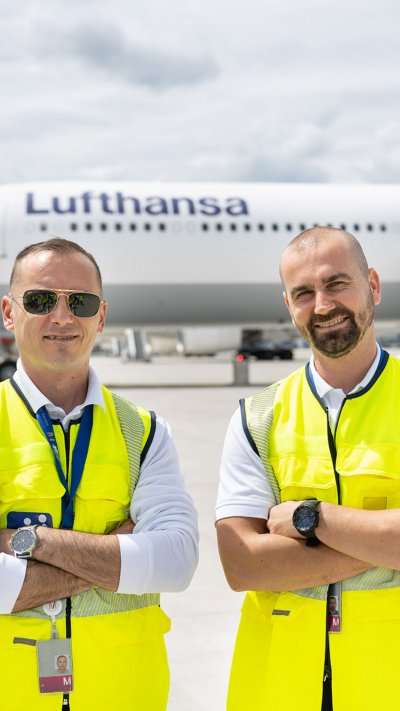 Zwei Service Agents stehen auf dem Vorfeld vor einer Lufthansa Maschine.