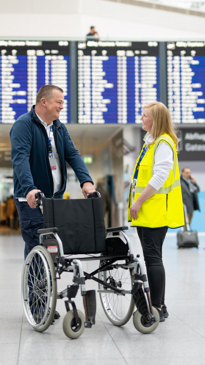 Zwei Servicemitarbeitende stehen in der Ankunftshalle des Flughafens neben einem Rollstuhl und unterhalten sich.