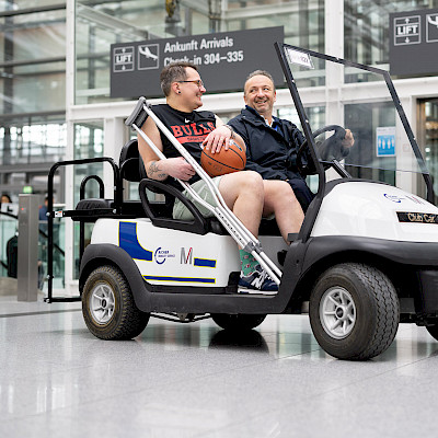 Ein verletzter Passagier in Sportkleidung fährt mit einem Service Agent im E-Car durch das Flughafengebäude.