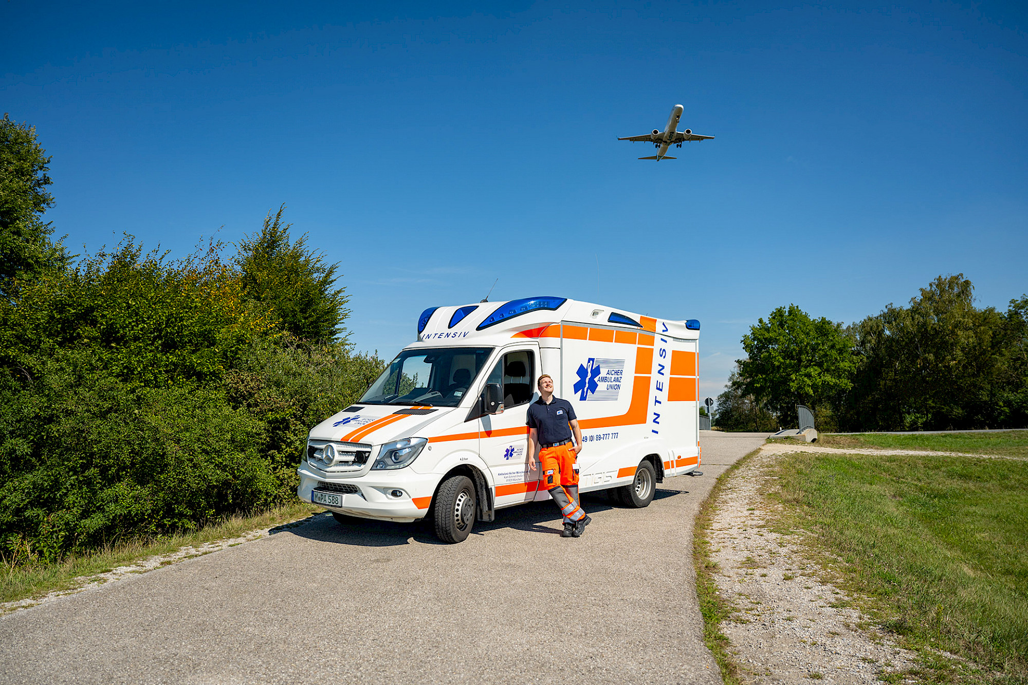 Ein Rettungssanitäter steht vor dem Rettungswagen, während im Hintergrund ein Flugzeug startet.