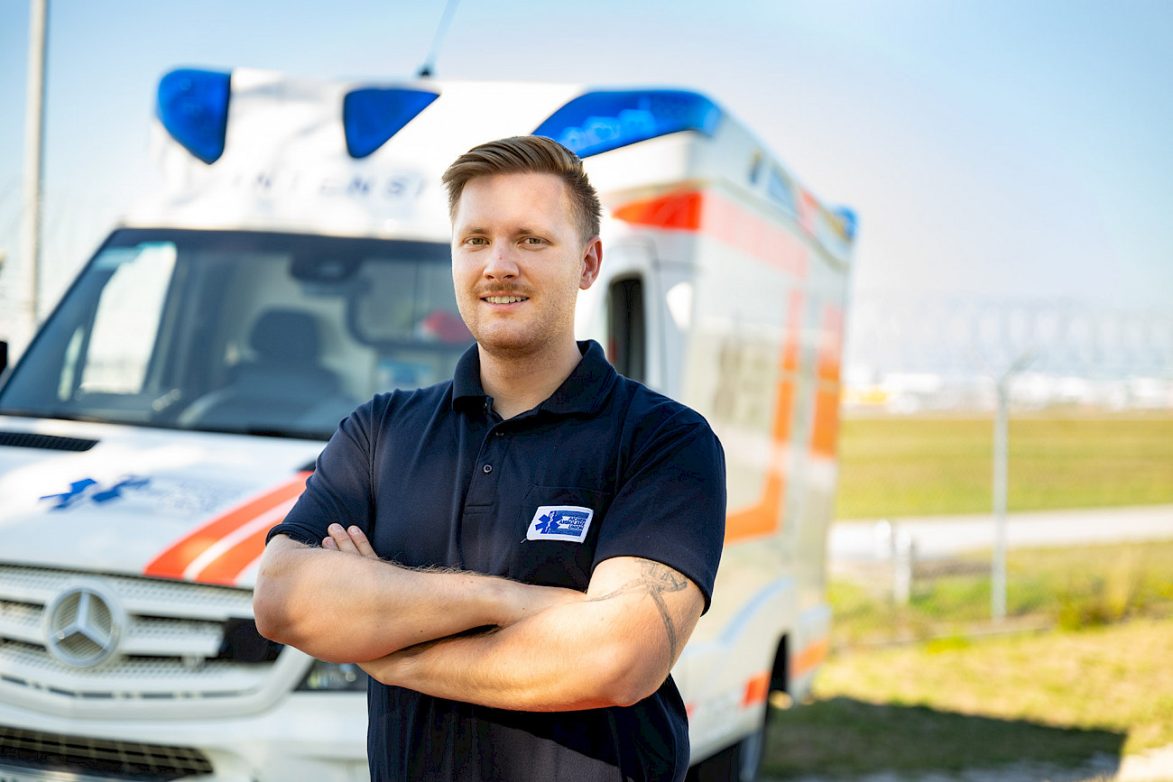 Freundlicher Sanitäter steht vor einem Einsatzwagen der Aicher Ambulanz Union..