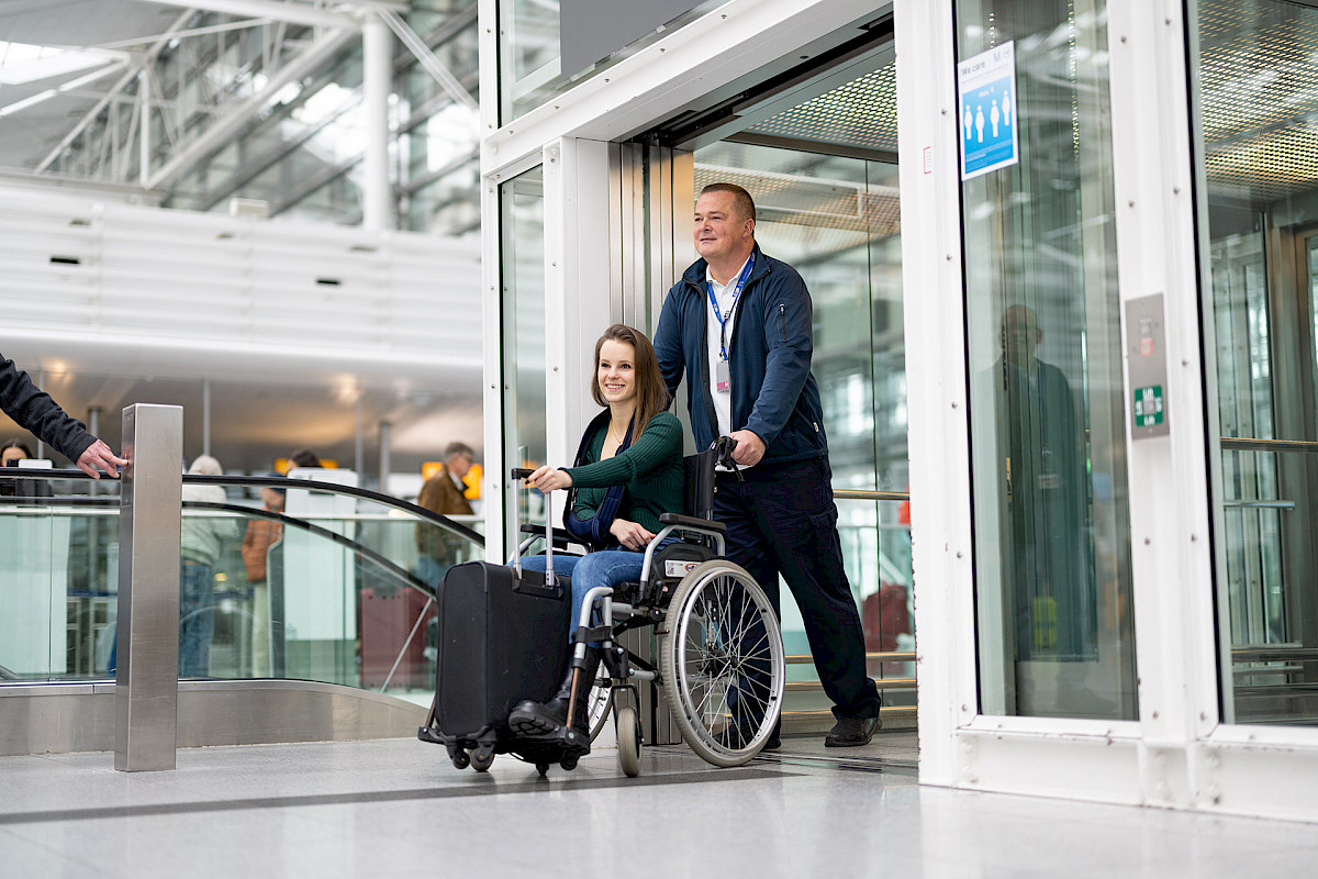 Service Agent schiebt Frau im Rollstuhl durch das Flughafengebäude.